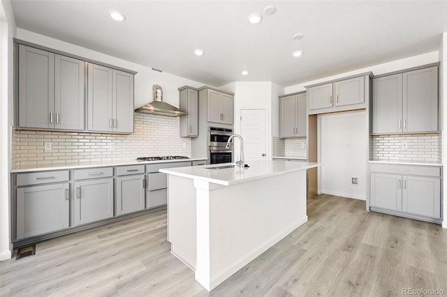 kitchen with wall chimney range hood, decorative backsplash, a kitchen island with sink, appliances with stainless steel finishes, and gray cabinetry