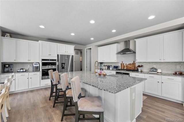 kitchen featuring light stone counters, a center island with sink, appliances with stainless steel finishes, white cabinetry, and wall chimney exhaust hood