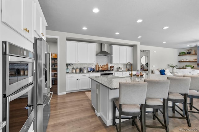 kitchen with a breakfast bar, white cabinetry, wall chimney range hood, appliances with stainless steel finishes, and a center island with sink