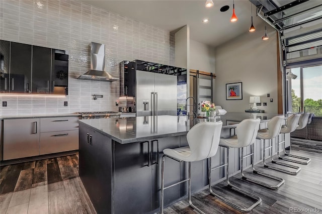 kitchen with high end fridge, dark wood-style flooring, a barn door, wall chimney exhaust hood, and tasteful backsplash