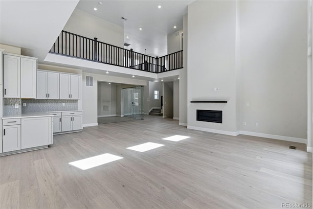 unfurnished living room with light wood-style flooring, visible vents, stairway, a towering ceiling, and a glass covered fireplace