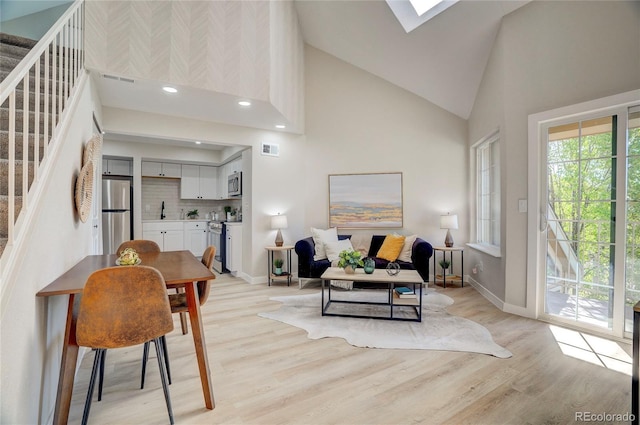 living room with a skylight, stairway, light wood-style floors, and high vaulted ceiling