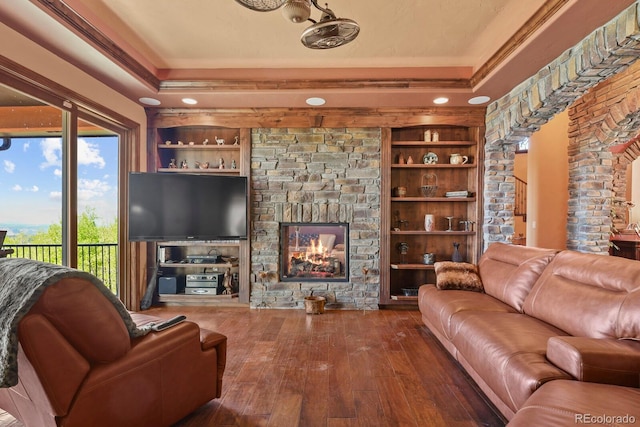 living room featuring ornamental molding, a tray ceiling, built in shelves, wood-type flooring, and a stone fireplace