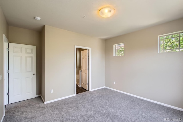 unfurnished bedroom with baseboards, multiple windows, ensuite bathroom, and dark colored carpet