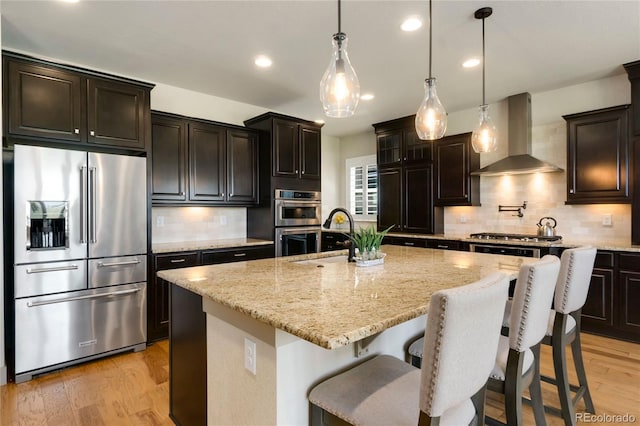 kitchen featuring wall chimney exhaust hood, light wood-style flooring, light stone counters, a sink, and stainless steel appliances