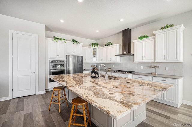 kitchen featuring wall chimney range hood, light wood finished floors, a sink, appliances with stainless steel finishes, and a kitchen breakfast bar