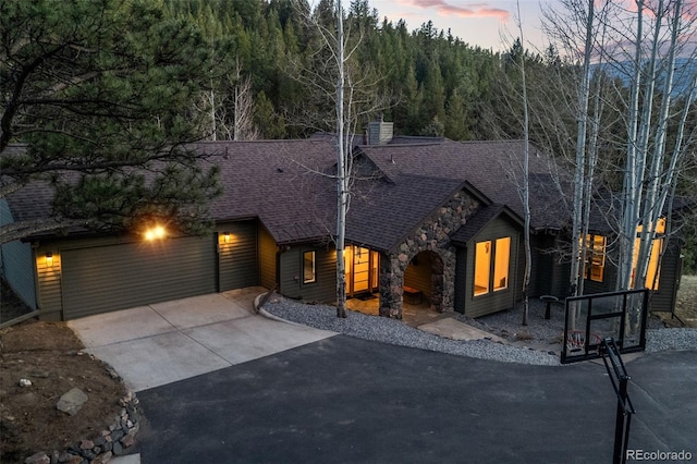 view of front of property featuring roof with shingles, stone siding, driveway, a view of trees, and a chimney