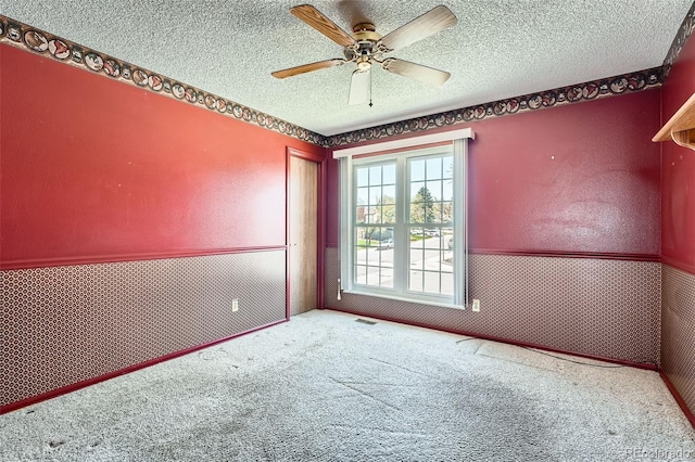empty room featuring visible vents, a textured ceiling, a wainscoted wall, a ceiling fan, and carpet