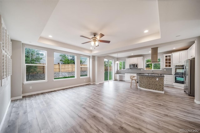 living area featuring a tray ceiling, ceiling fan, light wood-style flooring, baseboards, and healthy amount of natural light