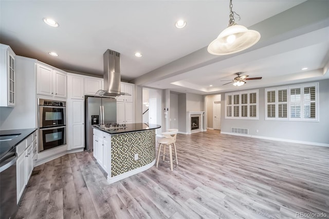 kitchen with island range hood, appliances with stainless steel finishes, a raised ceiling, dark countertops, and light wood-style floors