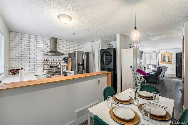 kitchen featuring wall chimney range hood, hanging light fixtures, stainless steel appliances, stacked washer and clothes dryer, and kitchen peninsula