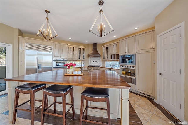 kitchen featuring stainless steel appliances, hanging light fixtures, cream cabinetry, custom range hood, and a kitchen island with sink