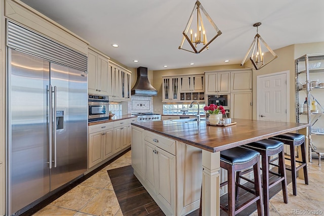 kitchen featuring wooden counters, custom range hood, cream cabinets, appliances with stainless steel finishes, and a center island with sink