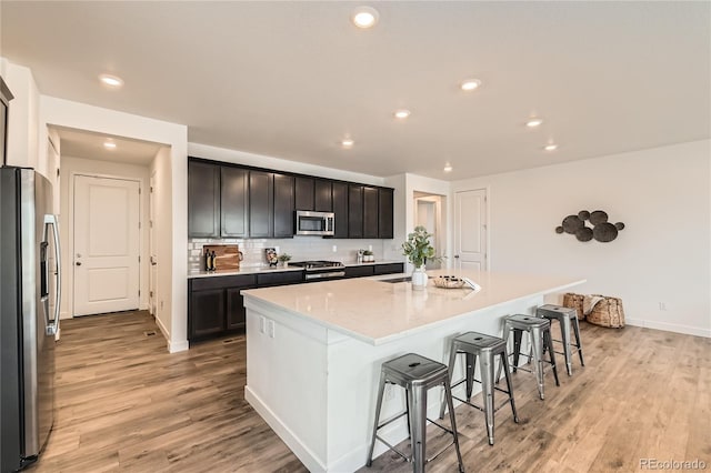 kitchen with a breakfast bar area, stainless steel appliances, light wood-style floors, backsplash, and an island with sink