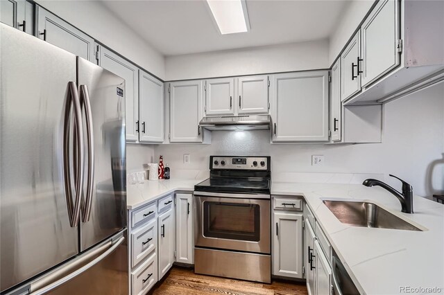 kitchen featuring light stone counters, under cabinet range hood, stainless steel appliances, wood finished floors, and a sink