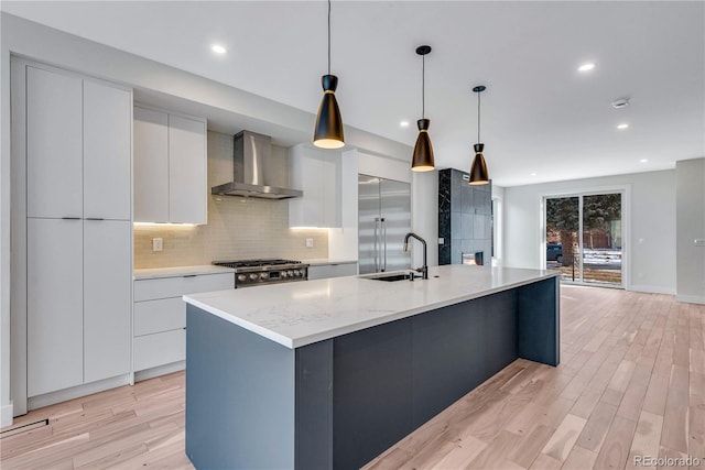 kitchen with wall chimney exhaust hood, sink, decorative light fixtures, a center island with sink, and white cabinets