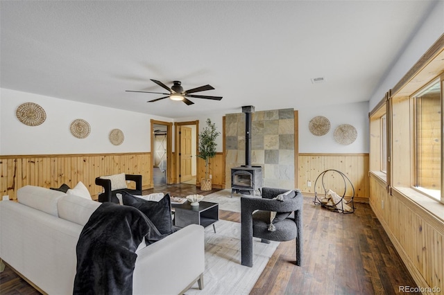 living area with wooden walls, a wood stove, a wainscoted wall, a ceiling fan, and hardwood / wood-style flooring