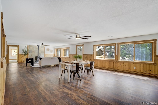 dining area with a ceiling fan, wainscoting, a wood stove, wood walls, and dark wood-type flooring