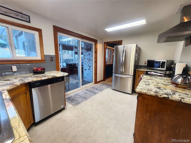 kitchen with wall chimney range hood, decorative backsplash, light stone countertops, light colored carpet, and stainless steel appliances