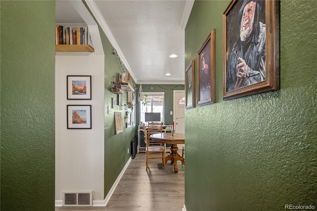 hallway featuring wood finished floors, baseboards, visible vents, ornamental molding, and a textured wall