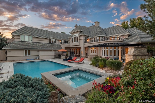 back house at dusk featuring a balcony, a swimming pool with hot tub, and a patio area