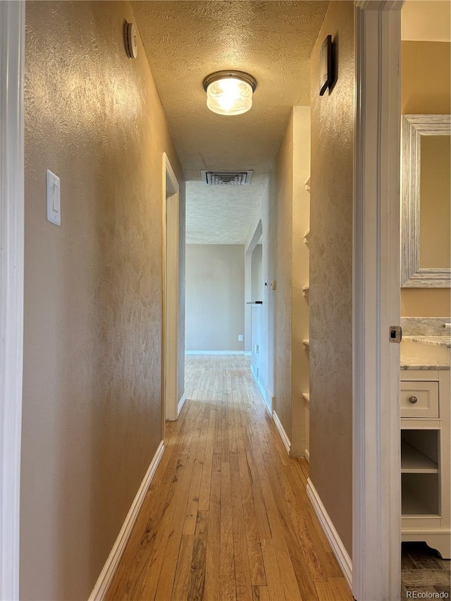 corridor with light wood finished floors, visible vents, a textured wall, a textured ceiling, and baseboards