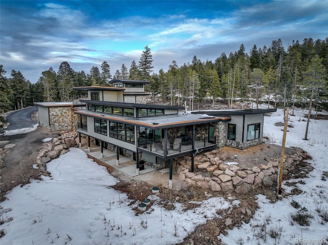 snow covered back of property with stone siding and a sunroom