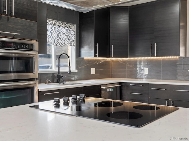kitchen featuring appliances with stainless steel finishes, a sink, dark cabinets, backsplash, and light stone counters