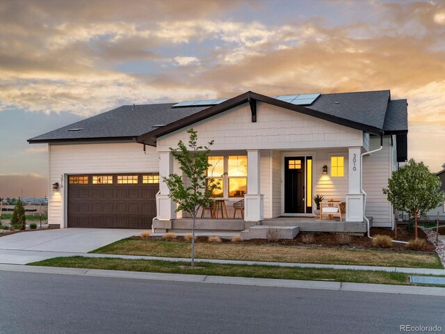 view of front facade featuring a shingled roof, a garage, solar panels, covered porch, and driveway