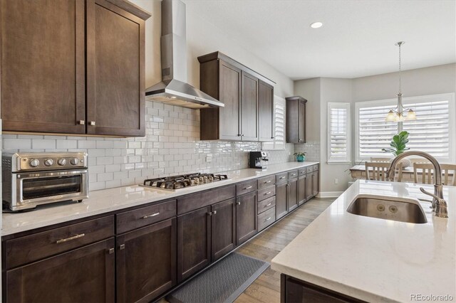 kitchen featuring wall chimney range hood, a sink, stainless steel gas cooktop, light wood-type flooring, and a chandelier