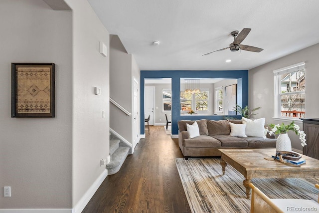 living area featuring stairs, ceiling fan with notable chandelier, baseboards, and dark wood finished floors