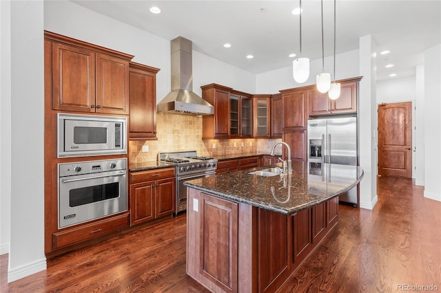 kitchen featuring a sink, wall chimney range hood, high end appliances, glass insert cabinets, and an island with sink