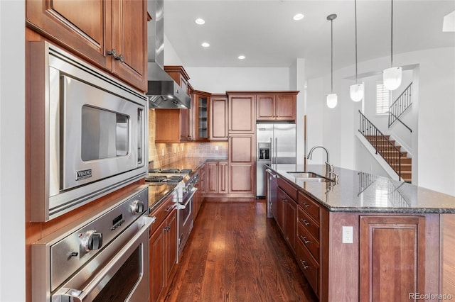 kitchen featuring glass insert cabinets, built in appliances, a kitchen island with sink, wall chimney range hood, and a sink