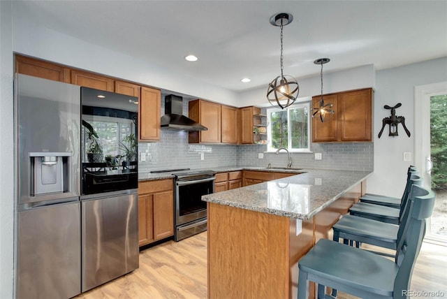kitchen with a peninsula, wall chimney range hood, brown cabinets, light wood-style flooring, and stainless steel appliances