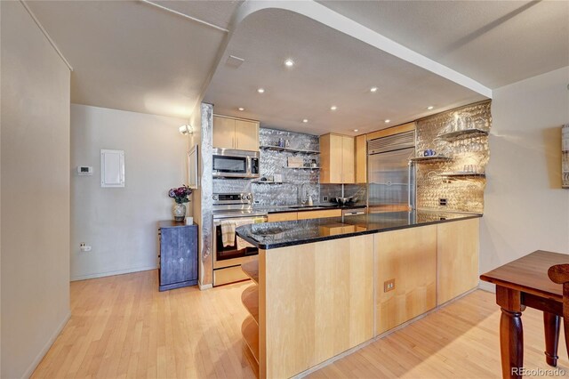 kitchen with kitchen peninsula, light wood-type flooring, dark stone countertops, backsplash, and appliances with stainless steel finishes