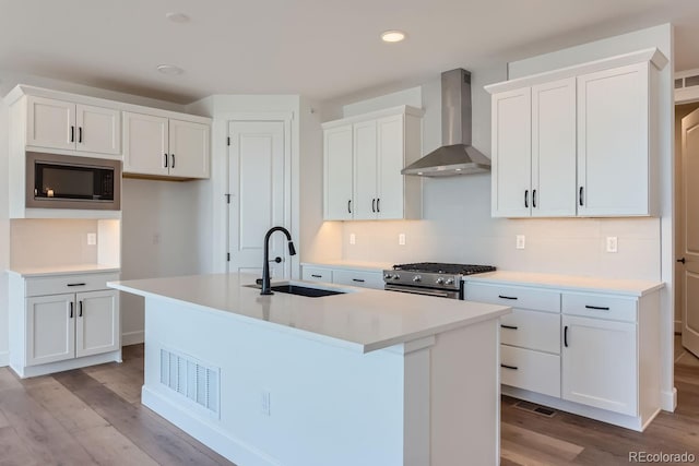 kitchen featuring a kitchen island with sink, white cabinets, wall chimney range hood, sink, and stainless steel gas range