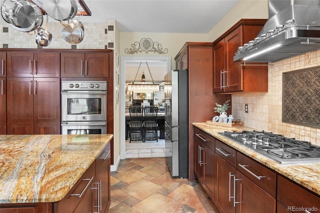 kitchen featuring wall chimney exhaust hood, appliances with stainless steel finishes, dark brown cabinets, tasteful backsplash, and light stone countertops