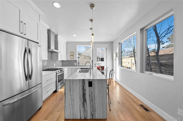 kitchen with white cabinetry, stainless steel appliances, a kitchen island with sink, hanging light fixtures, and wall chimney range hood