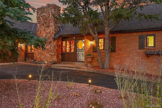 exterior entry at dusk featuring roof with shingles, a chimney, and brick siding