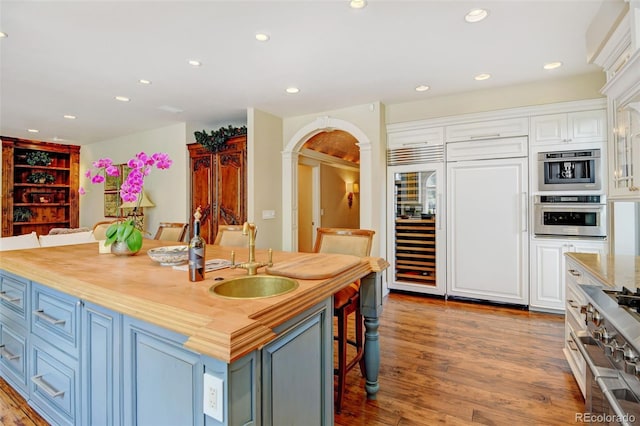 kitchen featuring arched walkways, a center island with sink, butcher block counters, white cabinetry, and a sink