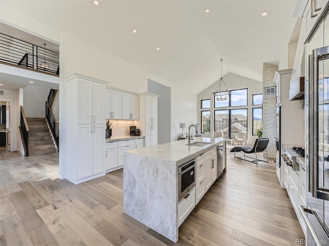 kitchen featuring high vaulted ceiling, light hardwood / wood-style floors, an island with sink, light stone counters, and sink