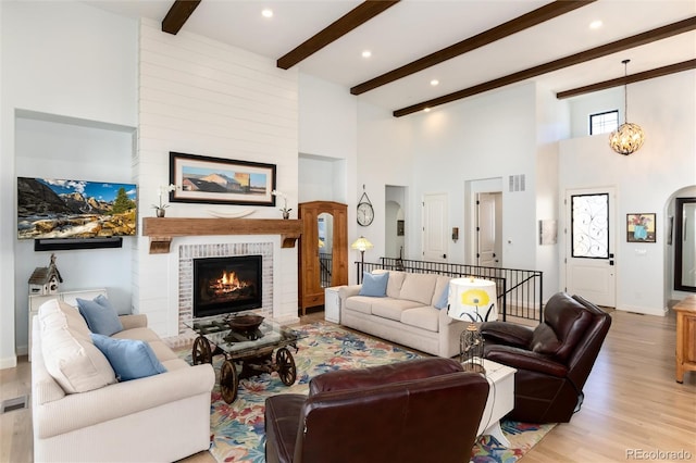 living room featuring a high ceiling, a brick fireplace, light hardwood / wood-style flooring, beamed ceiling, and a notable chandelier