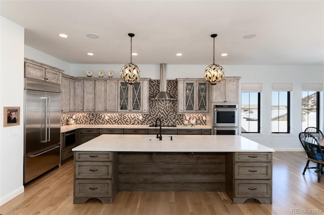 kitchen with built in appliances, wall chimney exhaust hood, an island with sink, and decorative light fixtures