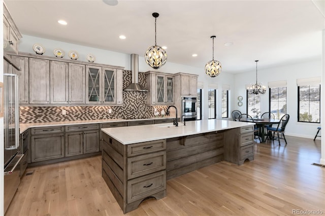 kitchen featuring wall chimney exhaust hood, an island with sink, decorative light fixtures, and light wood-type flooring