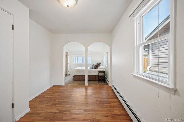 bedroom with arched walkways, wood finished floors, a baseboard heating unit, and baseboards