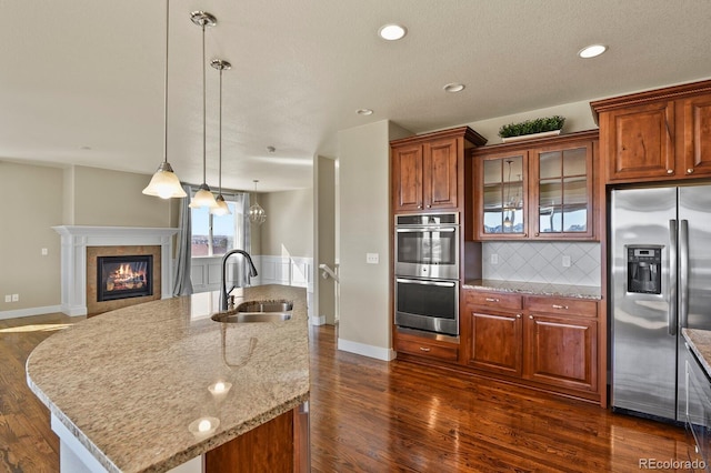 kitchen with an island with sink, stainless steel appliances, sink, light stone counters, and pendant lighting