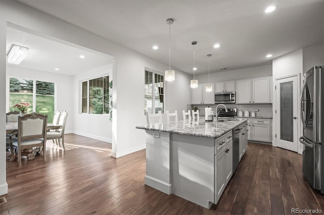 kitchen featuring stainless steel appliances, light stone countertops, dark wood-type flooring, a kitchen island with sink, and pendant lighting