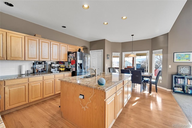 kitchen with sink, stainless steel refrigerator with ice dispenser, decorative light fixtures, light stone countertops, and a kitchen island with sink