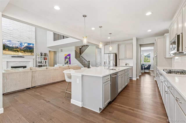 kitchen with pendant lighting, a center island with sink, a stone fireplace, light wood-type flooring, and stainless steel appliances