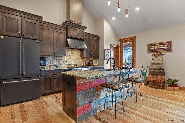 kitchen with dark brown cabinetry, under cabinet range hood, a breakfast bar, high end refrigerator, and a sink